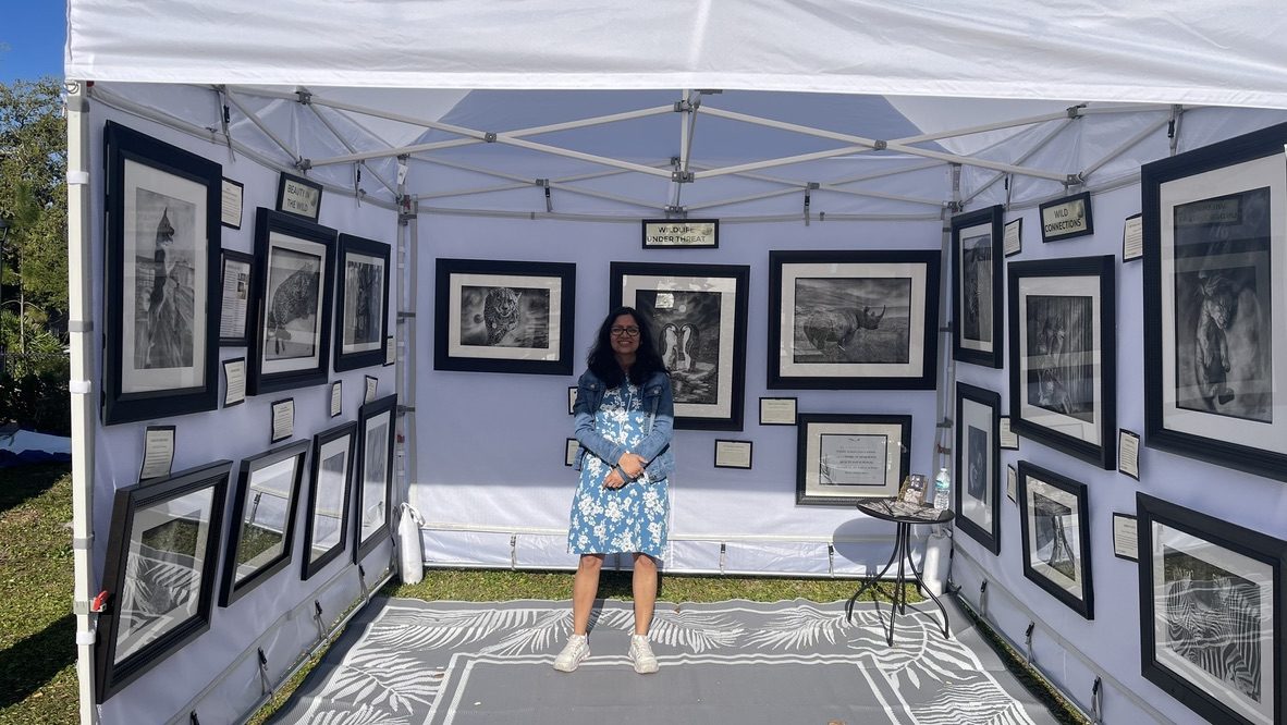 The artist in her booth at an outdoor park festival with framed drawings on both walls