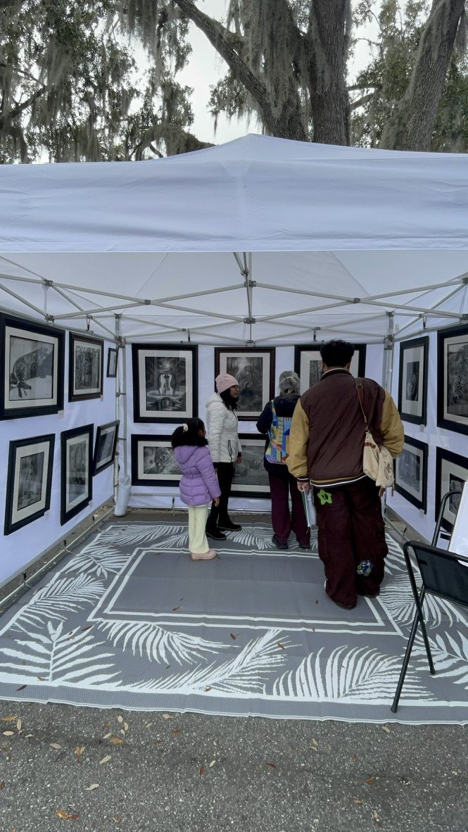 Art festival booth beneath live oaks draped with Spanish moss, visitors browsing framed wildlife drawings